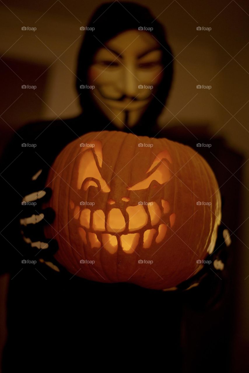 Man wearing Guy Fawkes mask holding spooky carved jack-o’-lantern pumpkin by @lynnebook