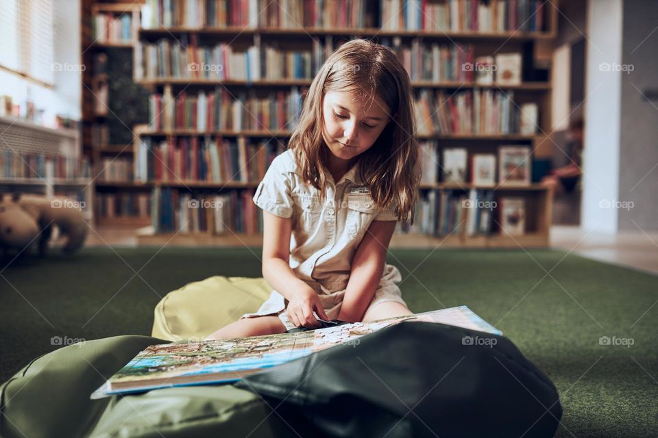 Schoolgirl doing puzzle and reading book in school library. Primary school pupil is involved in book with jigsaw. Smart girl learning from book. Benefits of everyday reading. Child curiosity