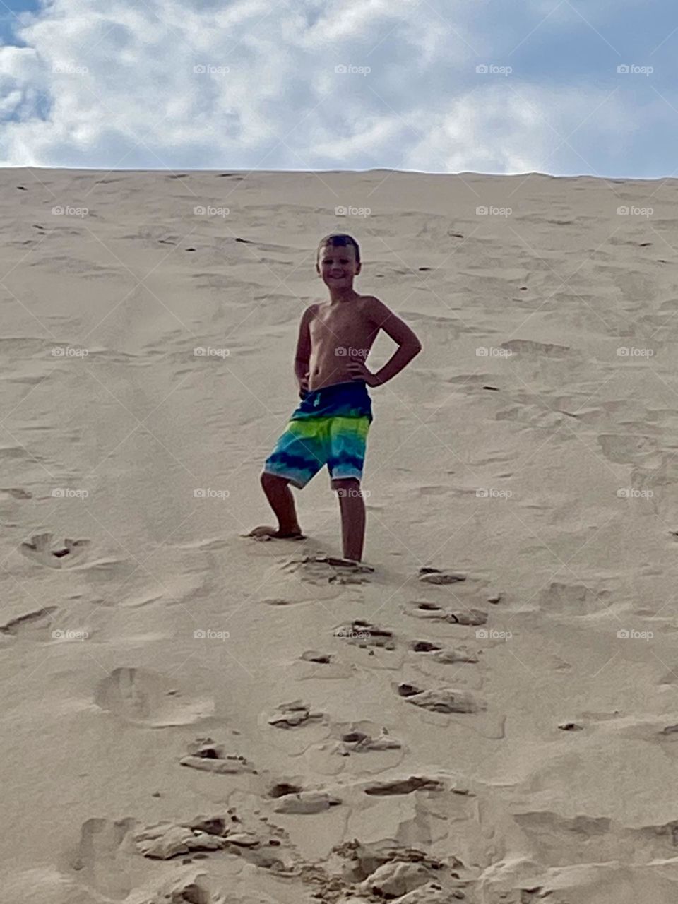 In this closer view, the person is more clearly visible standing on the sand dune, smiling and posing confidently. The bright, colorful swim shorts add a vibrant touch to the scene.