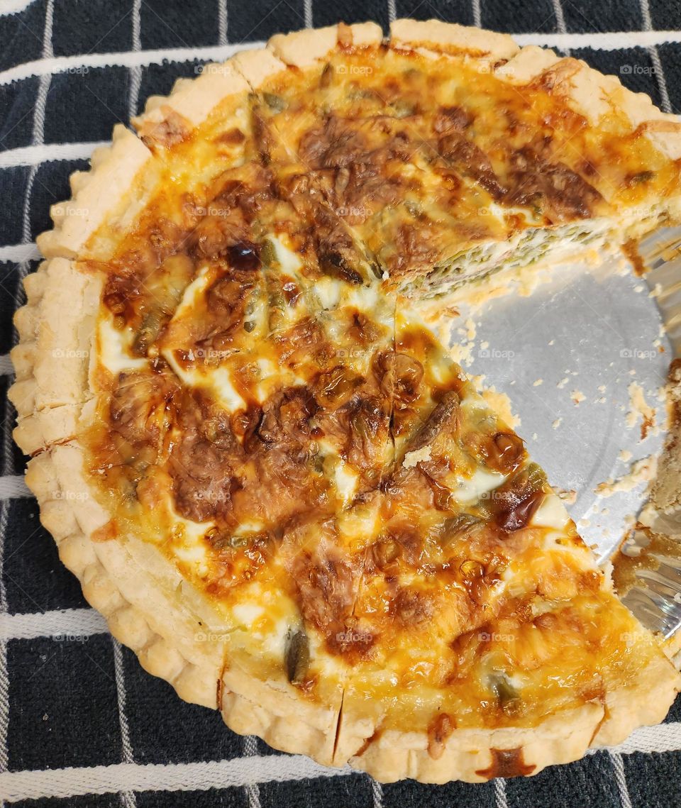 golden brown home baked quiche with slice removed from silver pie tin on black and white kitchen towel