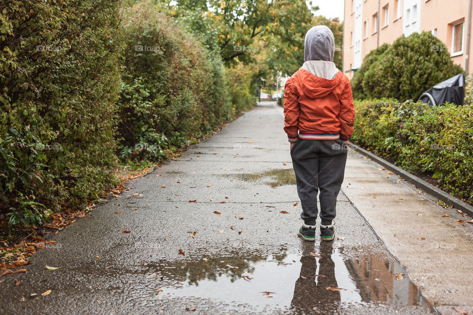 Boy standing puddle reflection. Preschool kid in autumn rain weather, mirror in water in urban environment