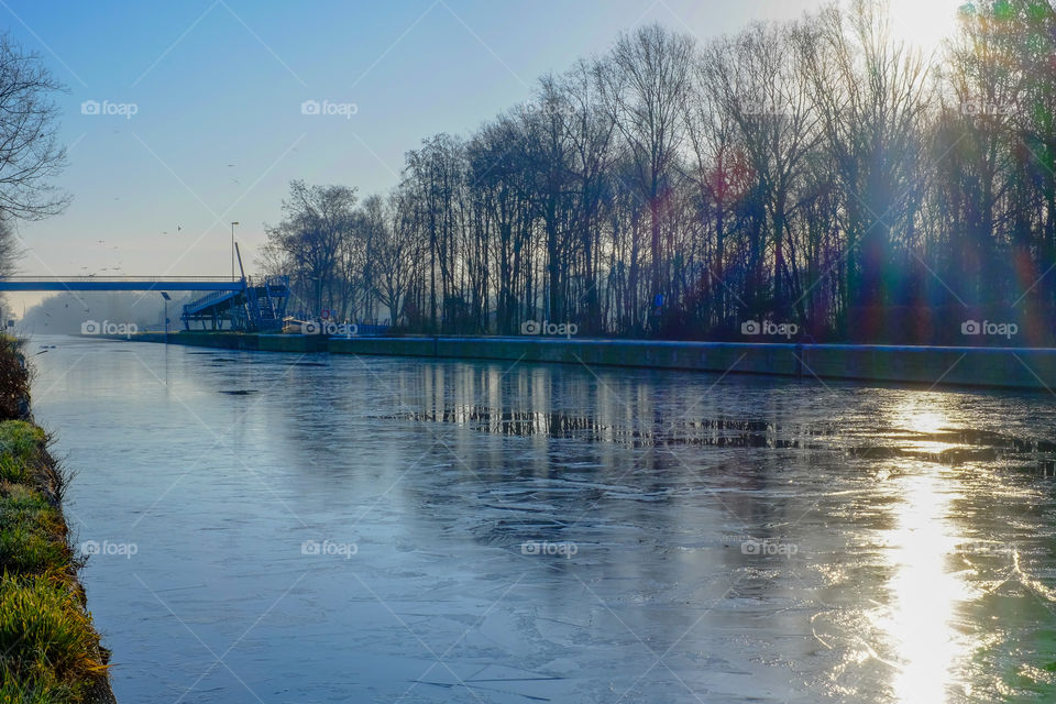 Winter sunrise over a frozen river running between the forest with the sun and the blue sky reflected on the ice and water