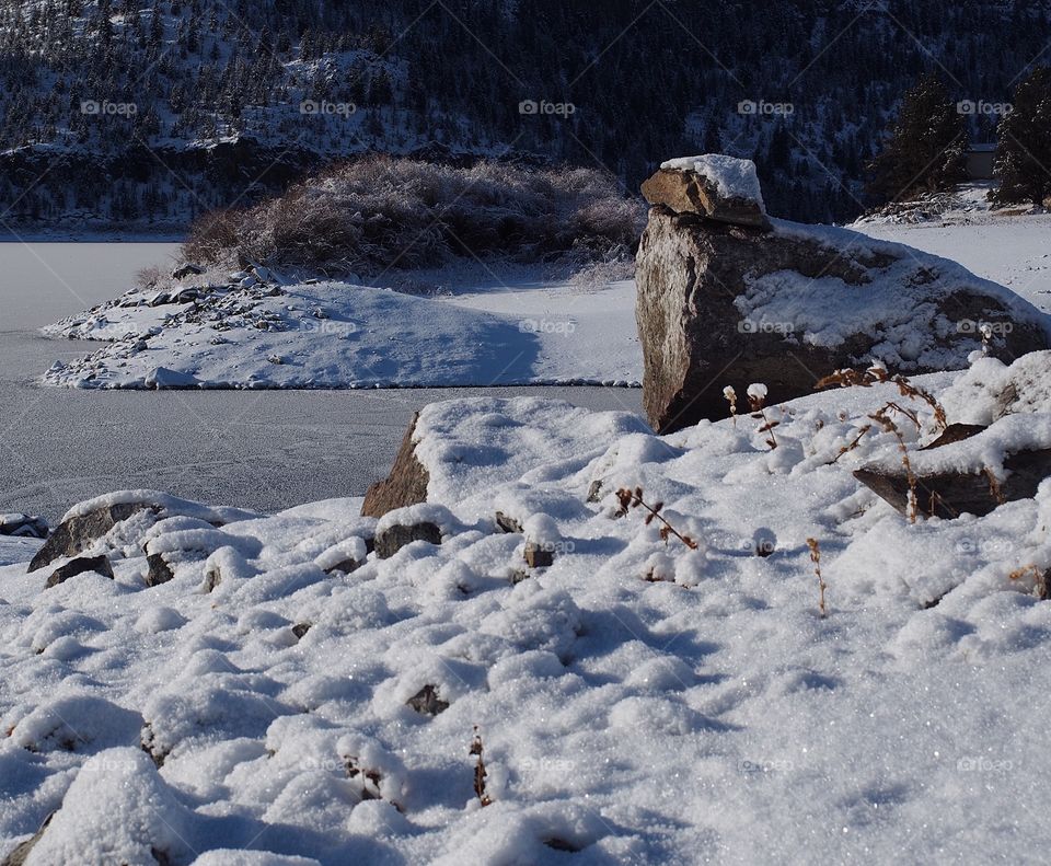 An inlet and a jagged boulder covered in snow at frozen over Ochoco Reservoir in Central Oregon on a freezing winter day. 