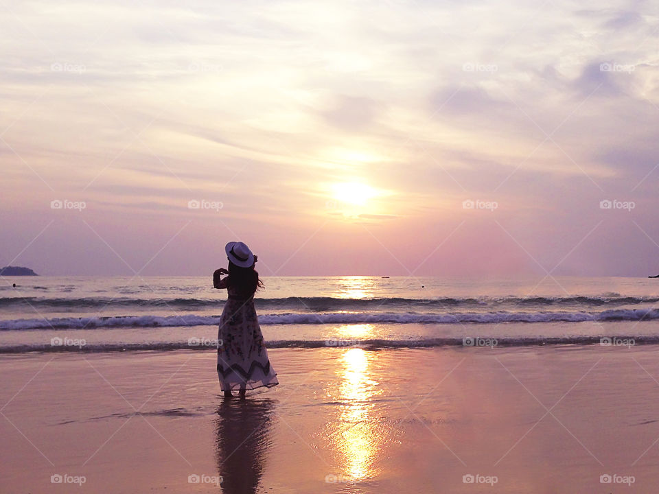 Silhouette of a young woman in summer dress with a fashionable hat in front of beautiful sunset above the beach 