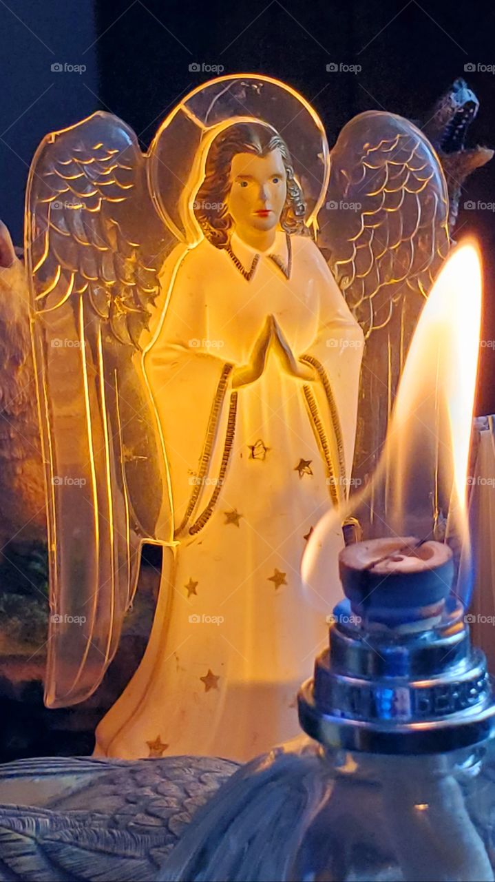 Burning flame on Lampe Berger wick filled with scented air cleansing oil. Vintage Angel nightlight lighted up standing in the background. Various other items are seen in the dim lighting.