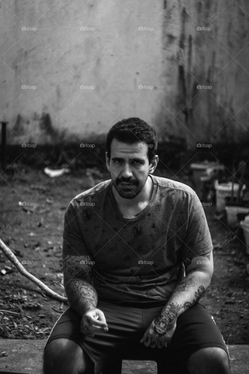 A Black and white portrait of a strong tattooed man, gazing at the camera, smoking a cigarette