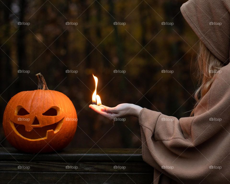 Cloaked woman reaching to light a carved pumpkin with a flame in her hand; fire magic