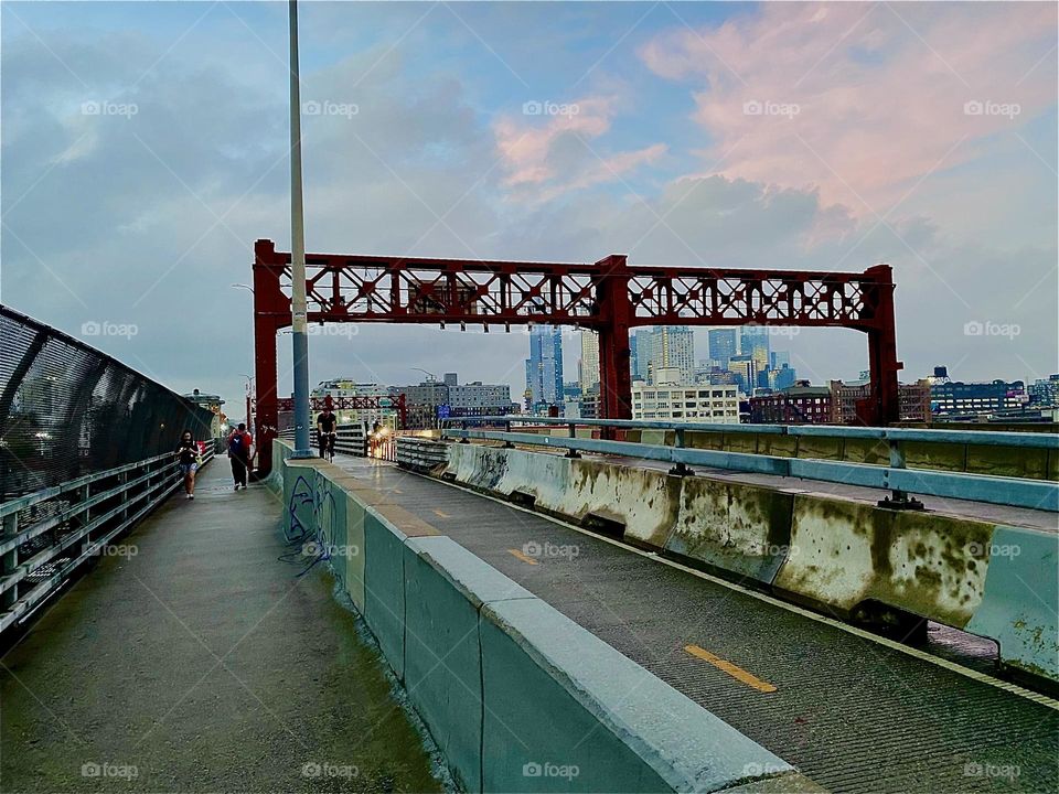 This is the pedestrian lane of the „Pulaski Bridge“ leading from „Greenpoint“, Brooklyn to LIC, Queens. It’s twilight time and the sun‘s slowly fading light is leaving imprints of pink on the still light blue sky. 2023. Hypnotic Productions