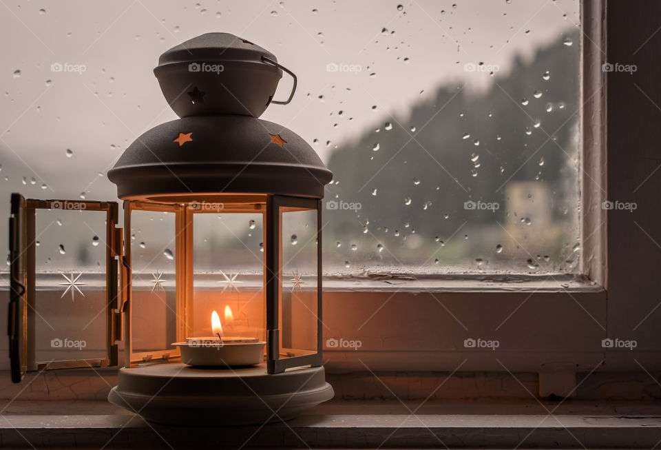 Opened lantern with lit candle in front of a window with raindrops
