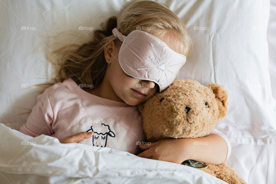 Cute little girl with blonde hair sleeping in bed with teddy bear toy