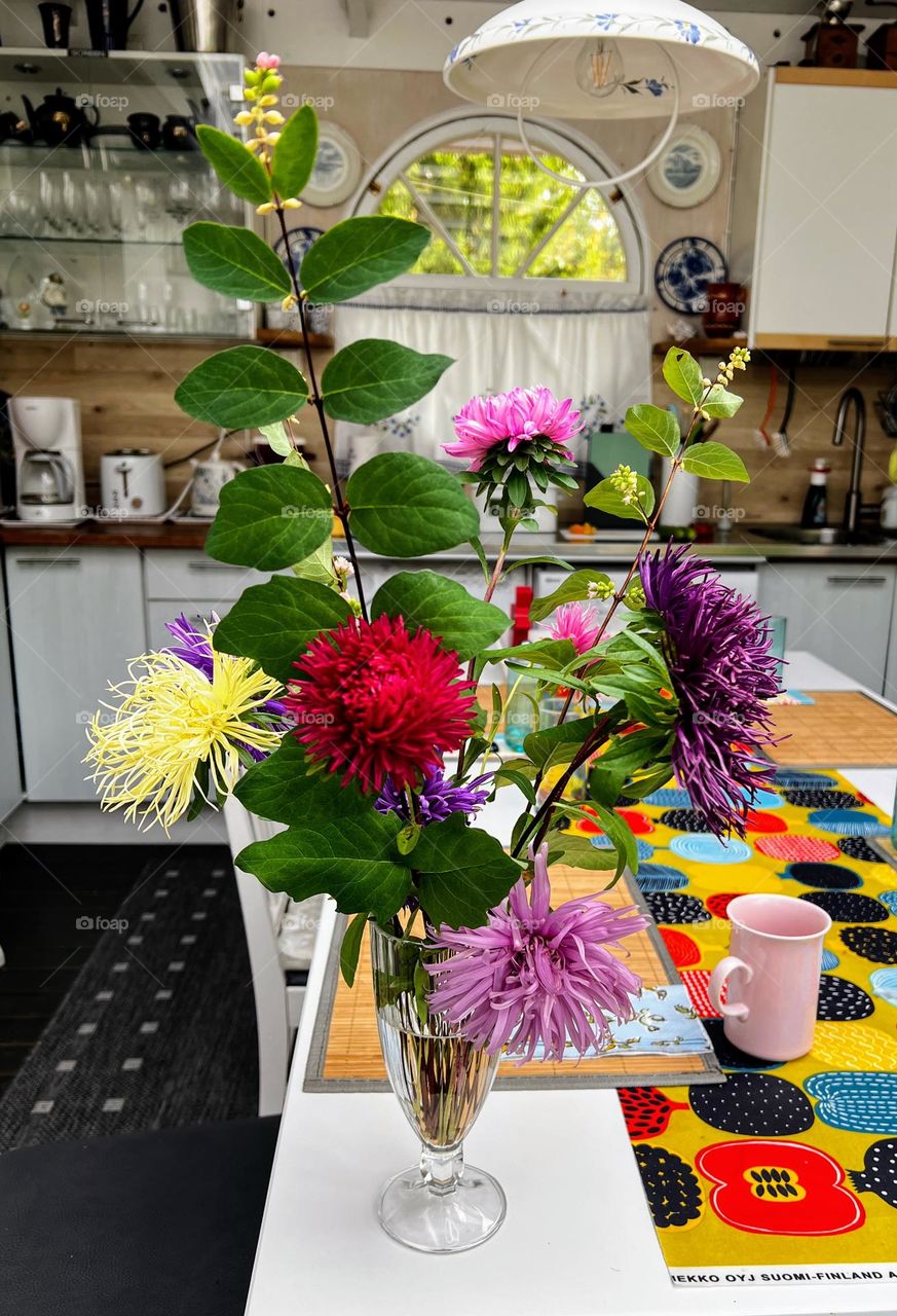 Beautiful multicoloured asters with some greens in the narrow vase on the table of the summer cottage 