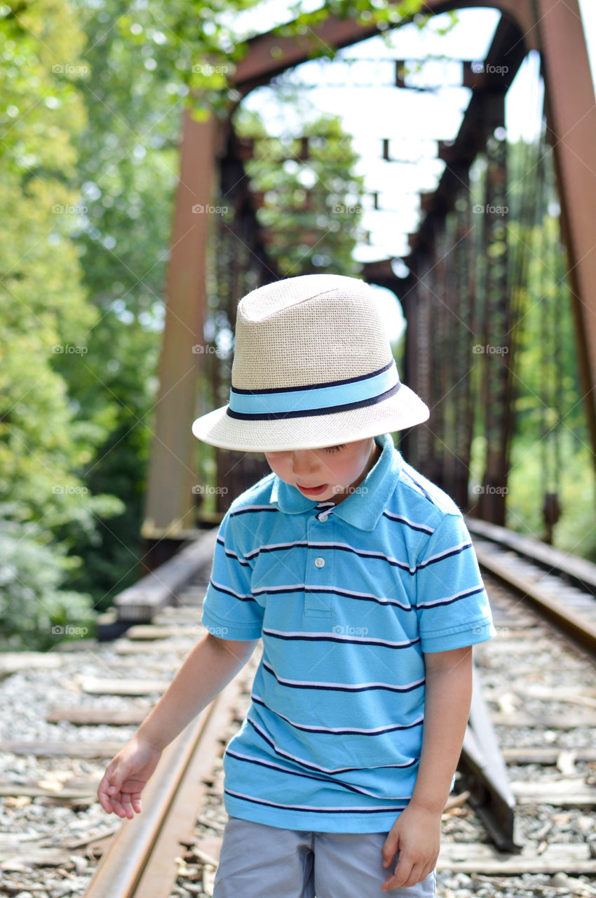 Young boy wearing a hat and looking down while walking along a railroad with a bridge in the background