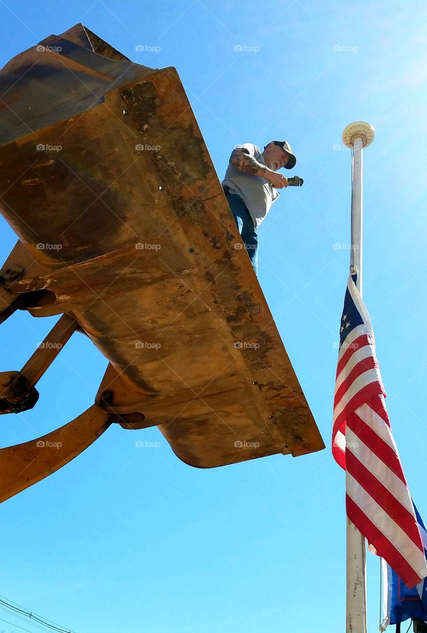 Man worker in bucket of front end loader. Dangerous reach to top of flagpole. Flag lowered 🇺🇸 to get at top. Worker reaching out. Dangerous work.