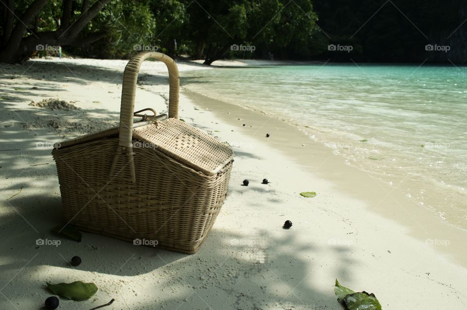 Beige picnic basket on the white sandy beach with azure sea and trees in the background