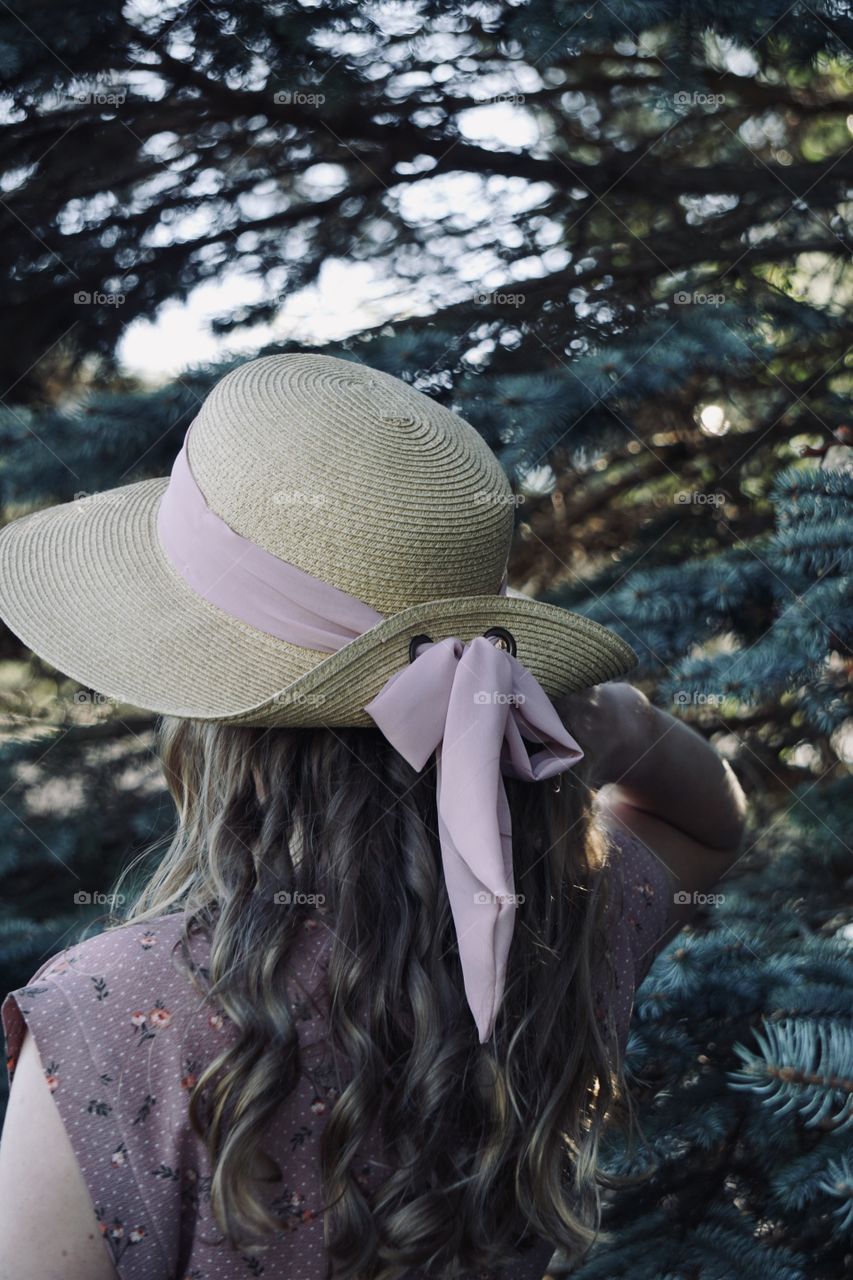 Girl wearing a hat, standing near some pine trees. 