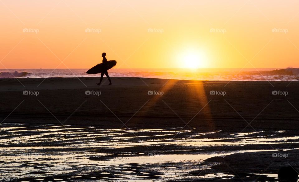 A surfer with board walks back across the beach at sunset