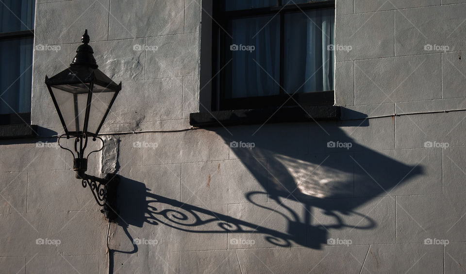 An street light, in the style of an old gas lamp casts a long an interesting shadow across a white building, underneath shutter windows