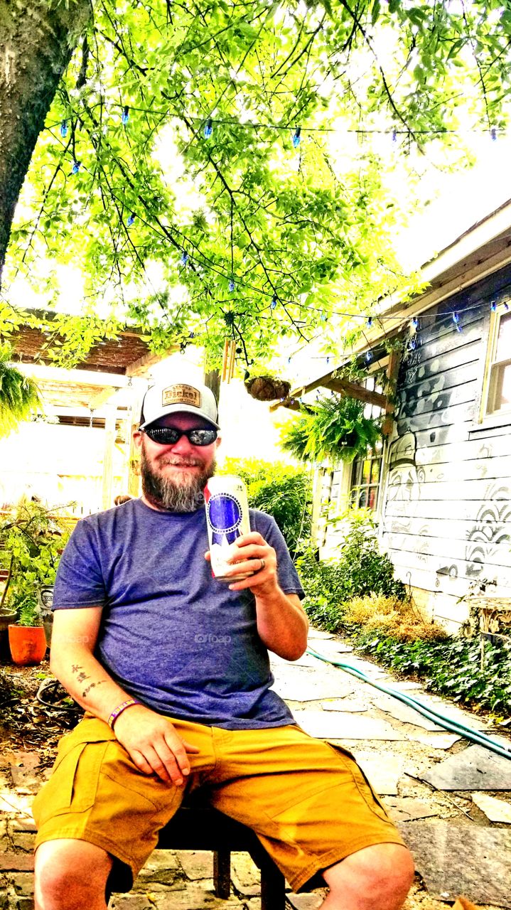 Summer day beer thirty for this smiley country guy, genuinely happy to cool off in the shade of the hovering green trees with an ice cold can of old school PBR on patio.