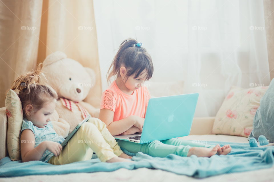 Little sisters with gadgets(laptop and tablet) in the bed.