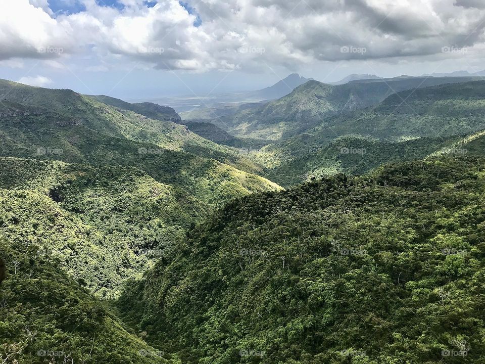 View across undulating hills in Mauritius under a cloudy sky causing dappled shade. Mountain in distance. Shades of green