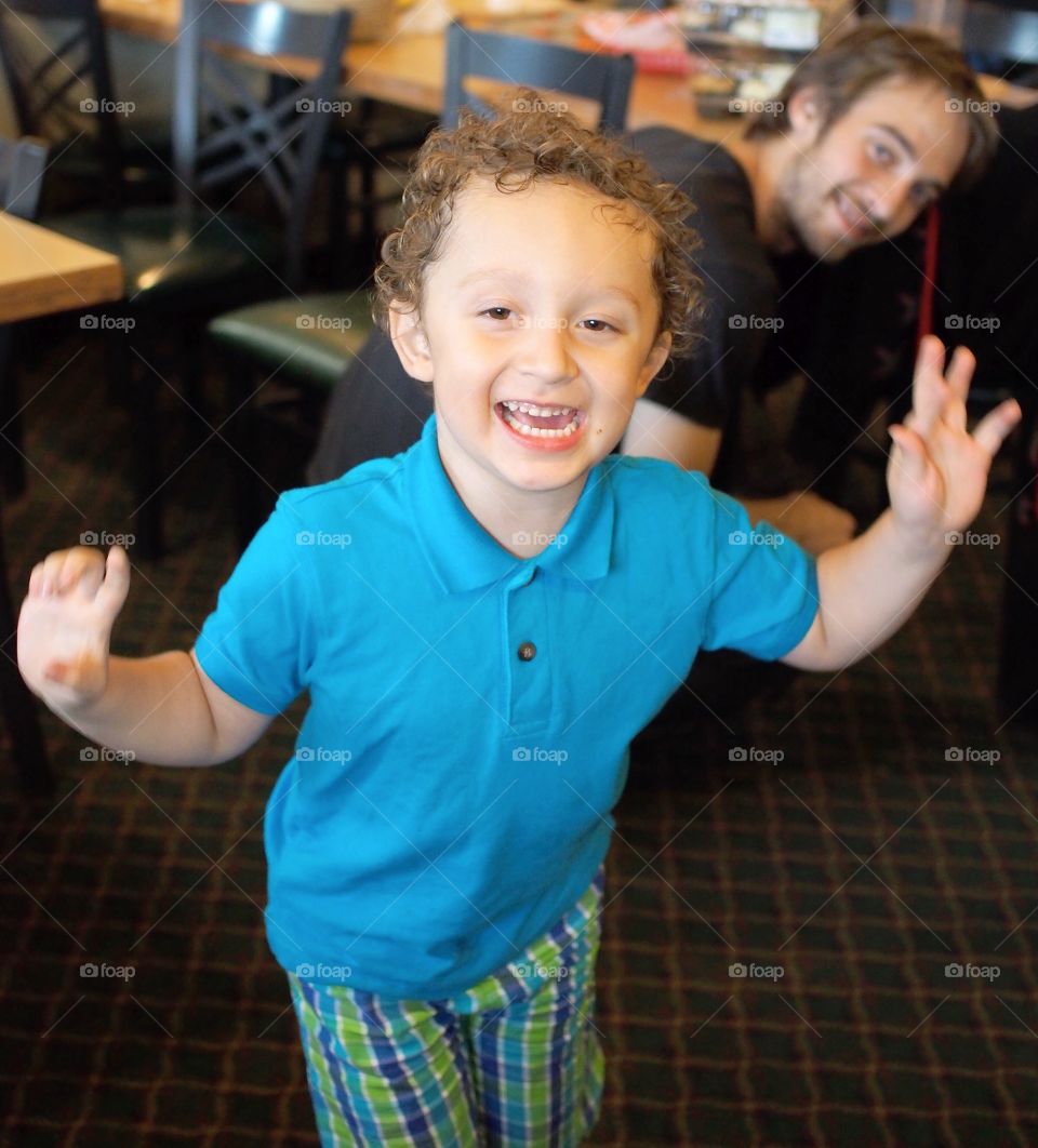 A excited young boy runs and plays at his indoor birthday party with his smiling father looking on in the background. 