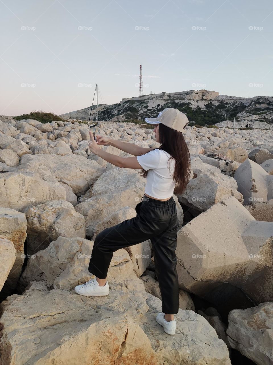 One young beautiful Caucasian brunette girl with flowing long hair in stylish clothes stands on a rocky cliff and takes a selfie video on a smartphone on a summer evening on the island of Frioul in France, close-up side view.