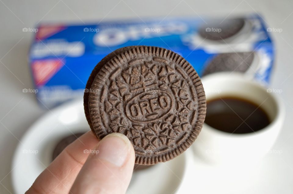 A persons fingers holding an Oreo cookie and a cup of coffee in the background