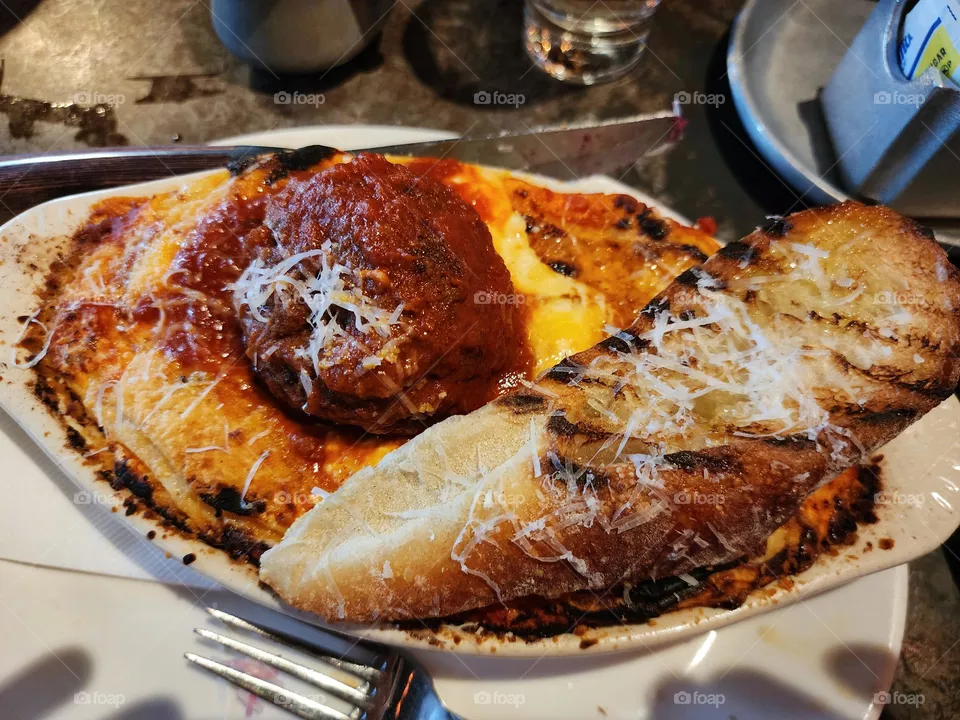 Baked lasagna with a meatball in the center and garlic bread off to the side.