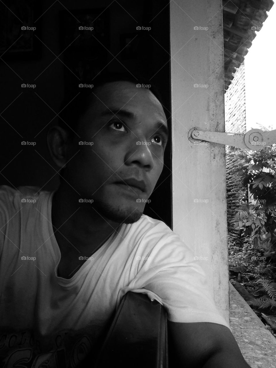 selfie photo of a man facing out of the room with his eyes looking up, photographed beside the window of the house in black and white style.