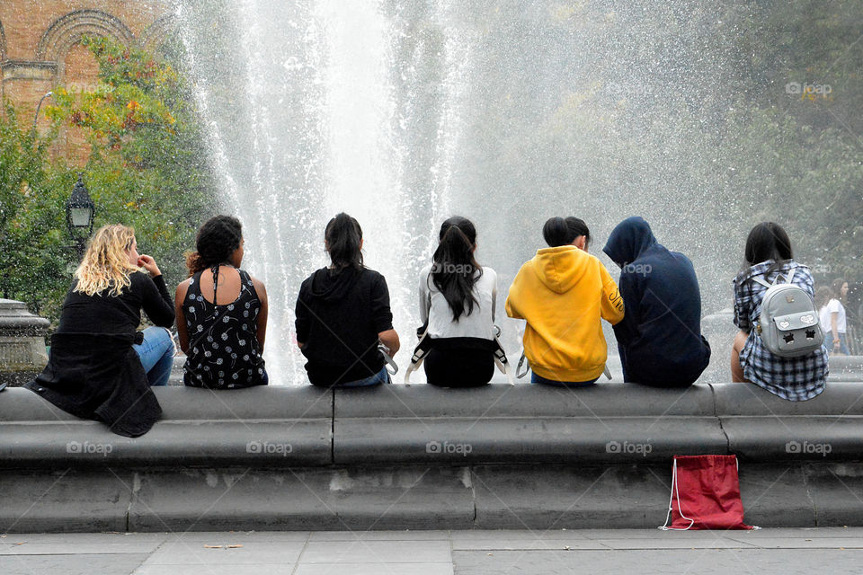 Unrecognizable people with their backs towards the camera sitting around a fountain in Washington Square park New York City.