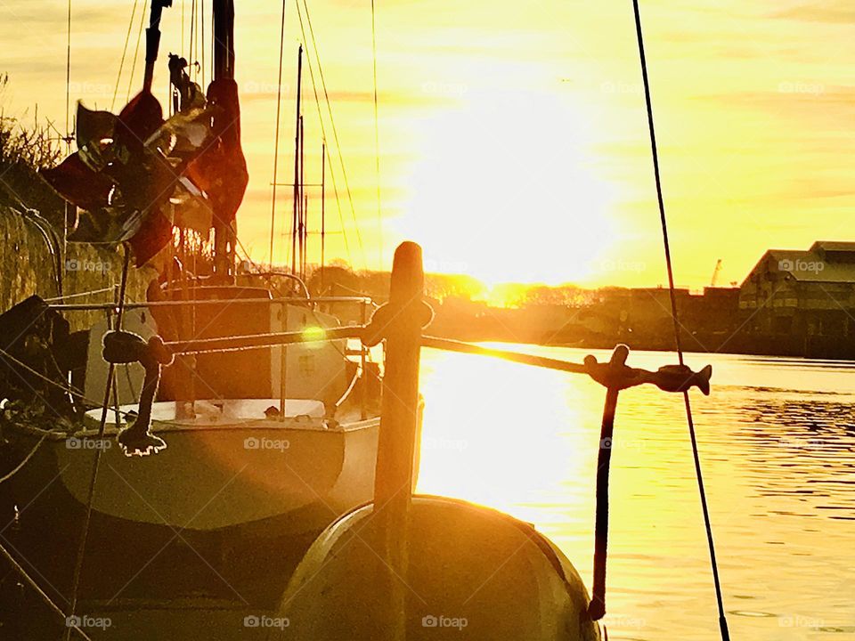A golden glowing sun setting slowly behind the horizon photographed from the shore at Newtown Creek, Long Island City, Queens, NY with a view of our sailboat “Salvation” in the foreground. 2018. Hypnotic Productions