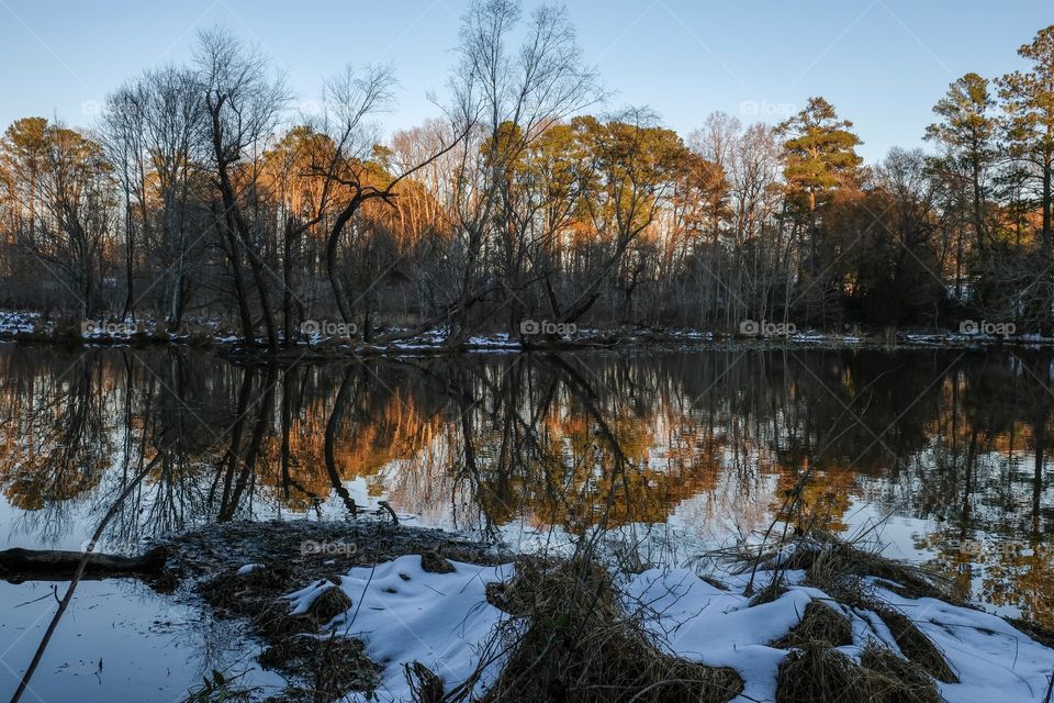 The last sliver of the day’s sunshine falls upon the treetops across the pond with mirror reflections on the still surface and patchy remnants of snow on the ground at Yates Mill County Park in Raleigh North Carolina. 