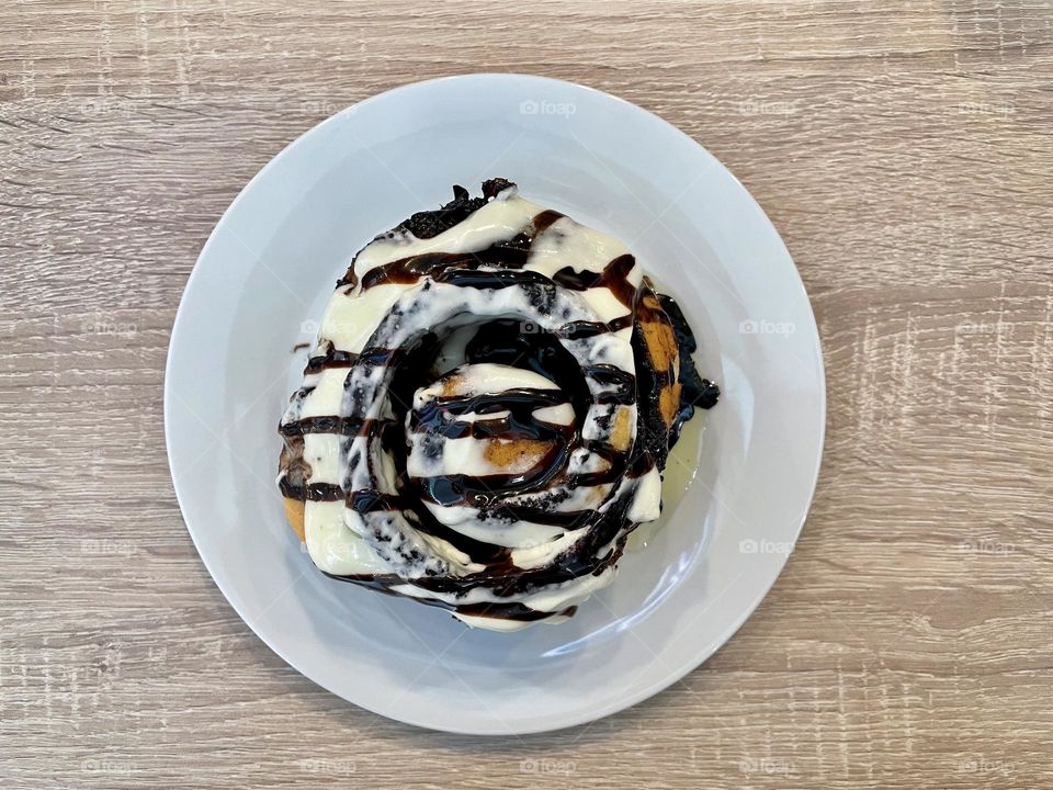 A big delicious cinnamon roll topped with white and dark chocolate sauce on a white plate on a wooden table.