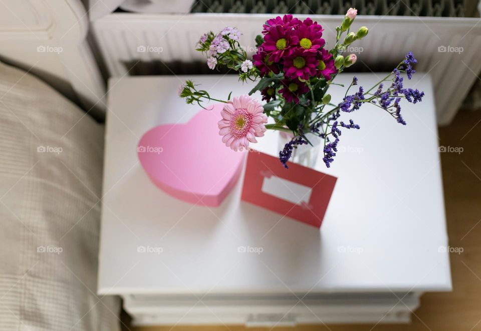 A beautiful view of a delicate bouquet of flowers in a vase, a pink heart box and an envelope stand on a white night table next to the bed, close-up. Concept romantic surprises, holidays.