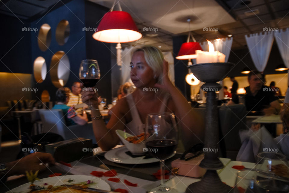 Un elegante restaurante con la mesa llena de vajilla y cristalería distinguida invita a acompañar a la mujer visualizada  en la foto a compartir el vino que está tomando .  El ambiente relajado y acogedor la envuelve con la sutil luz de vela .
