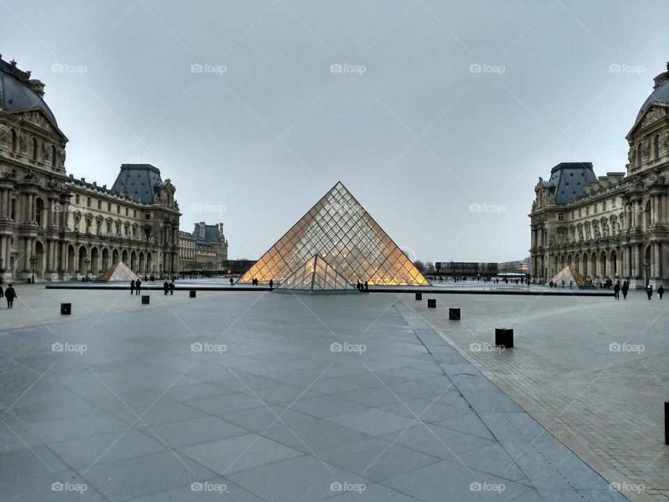 Exterior of the bright Louvre museum, on a tiled sidewalk and another cobblestone sidewalk, surrounding buildings and a white sky.