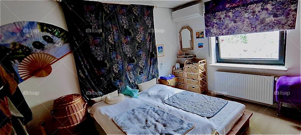 This is the master bedroom in the beautiful house in “Bavaria”, Germany. Wicker rattan furniture, a tapestry, a matching pull down curtain over the window to the garden, artwork, a mirror are part of the interior design. 2023. Hypnotic Productions