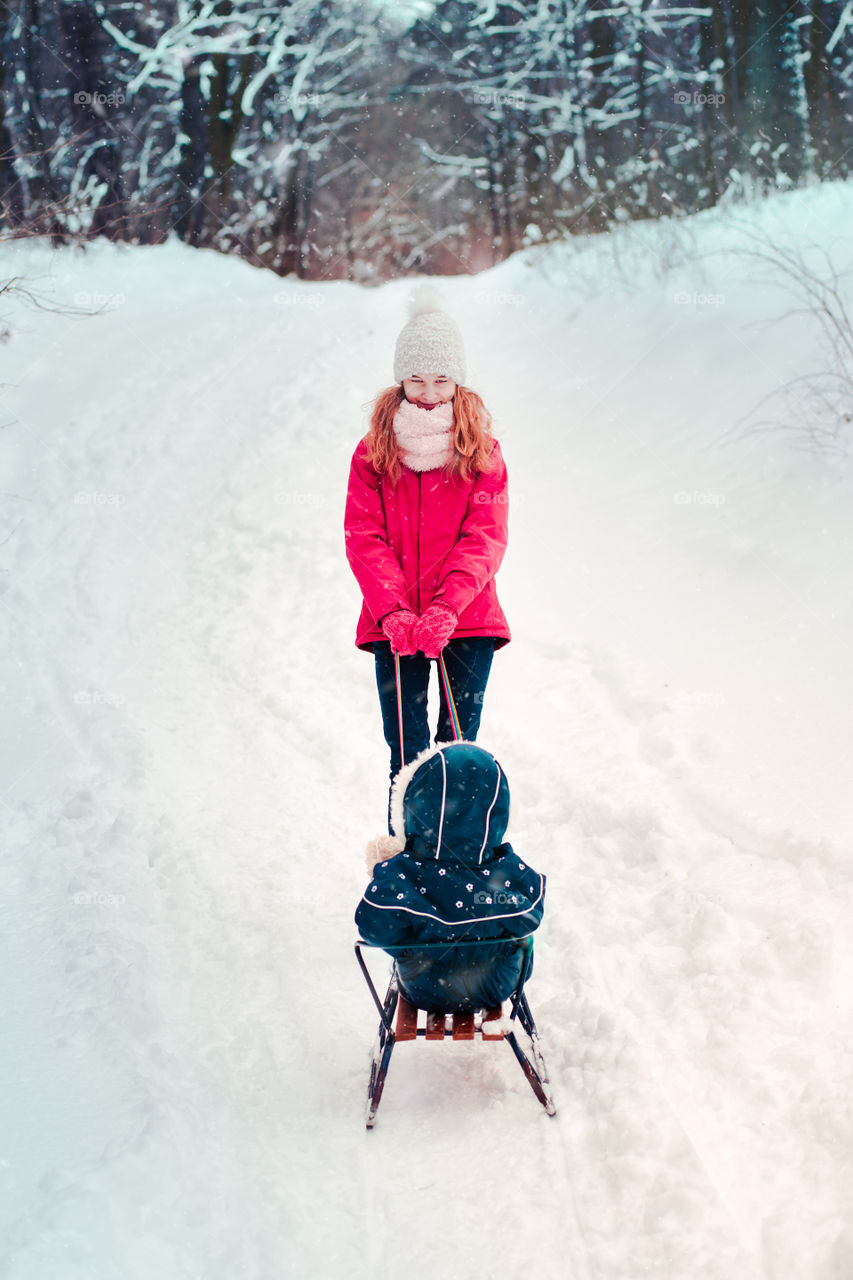 Teenage girl pulling sled with her little sister, a few years old girl, through forest covered by snow while snow falling, enjoying wintertime, spending time together. Girls are wearing winter clothes