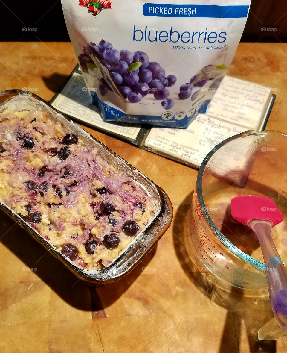 Blueberry Bread fixings show recipe, blueberries, glass mixing bowl, spatula. Bread ready to be baked in oven.