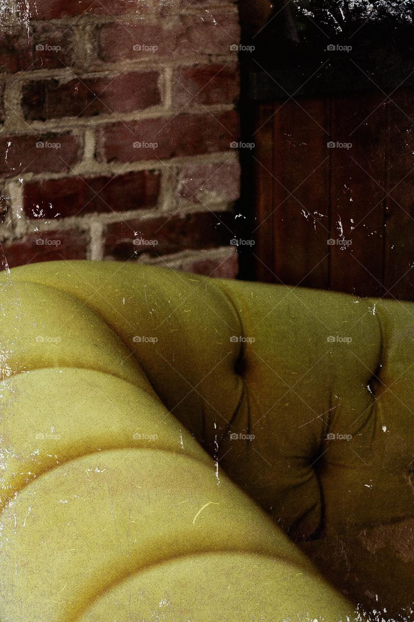 A closeup of a greenish-gold vintage styled sofa beside a wood panel and brick wall. Photo editing effects used to simulate an old worn vintage photo. 