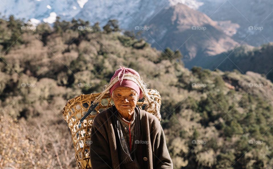 Old nepalese woman smiling, wearing her basket with a rope attached to the head and standing in front of a mountain background.