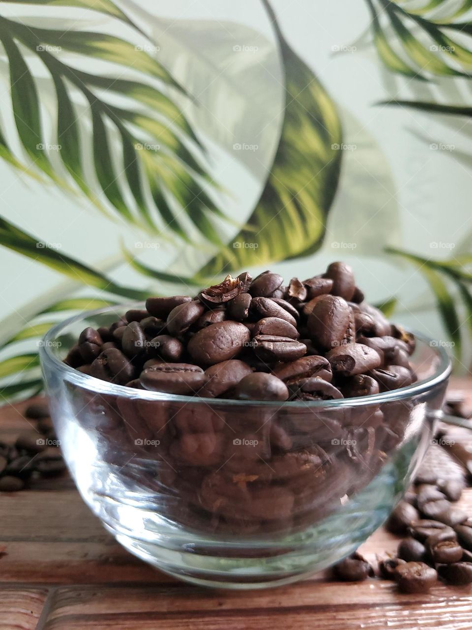 Coffee beans in a clear glass bowl on a wood surface with a green leaf wall paper background.