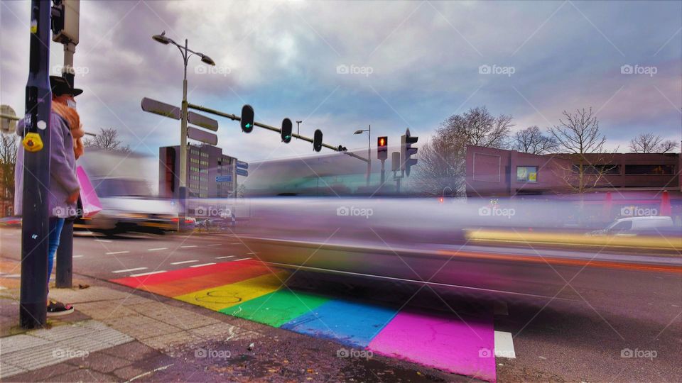 rainbow lbgtq pedestrian crossing with traffic lights and a waiting pedestrian with hat and a white car passing at full speed