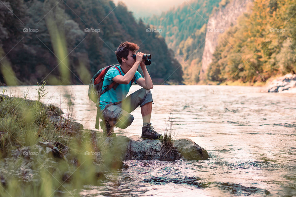 Young tourist with backpack looks through a binoculars on mountains peaks, stands on a rock over a river. Boy spends a vacation in mountains, wandering with backpack, he is wearing sports summer clothes