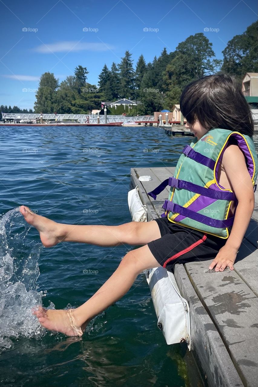 A young child kicks their feet in the water of a clear blue lake, splashing at a dock’s edge