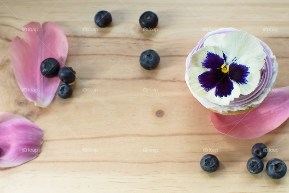 Floral rose and cupcake background with lavender and vanilla frosting cupcake with pansy on top and fresh scattered blueberries on wood desktop 