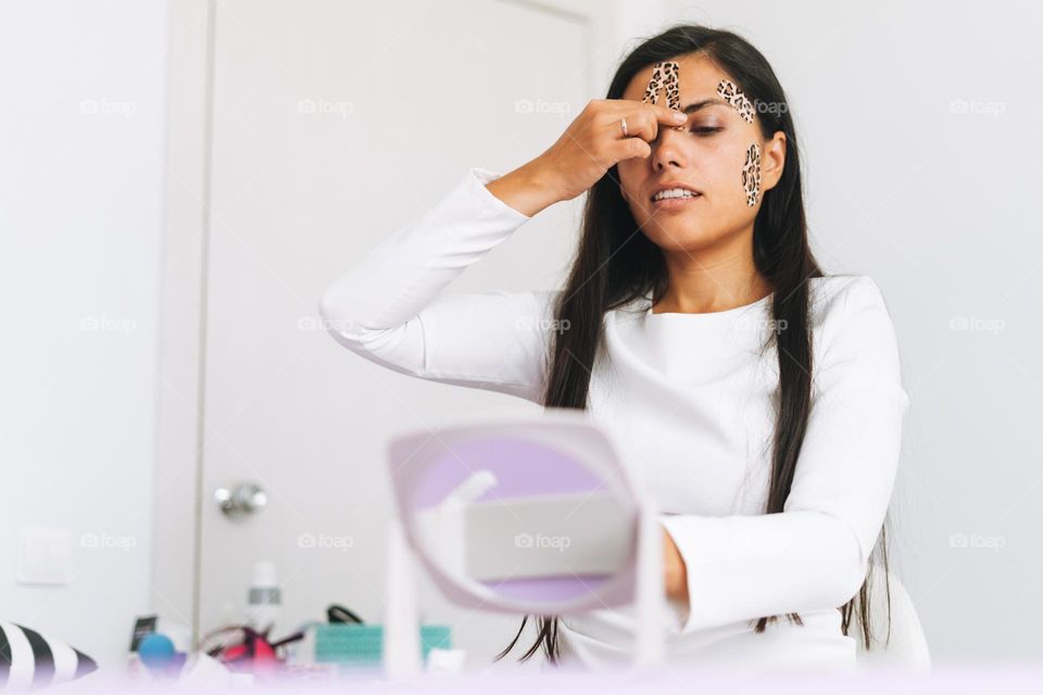 Young beautiful brunette woman with long hair cosmetologist makes face taping looks in the mirror in office. Young girl does anti-aging procedures with help of kinesio tape