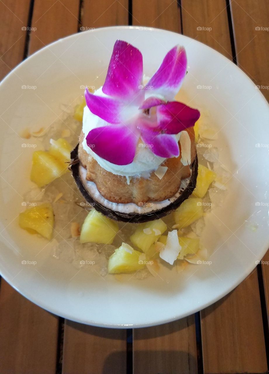 Delicious, coconut cake served in a coconut, topped with fresh pineapple ice cream. Decorated with pineapple cubes, coconut shavings, and a beautiful flower.