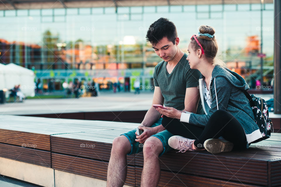 Couple of friends, teenage girl and boy,  having fun together, using smartphones,  sitting in center of town, spending time together. Real people, authentic situations