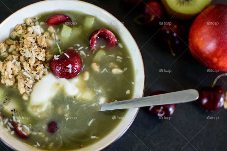 Eating a sweet Cherry and kiwi green smoothie bowl still life with coconut, chia and granola on counter with fresh cut fruit in background 